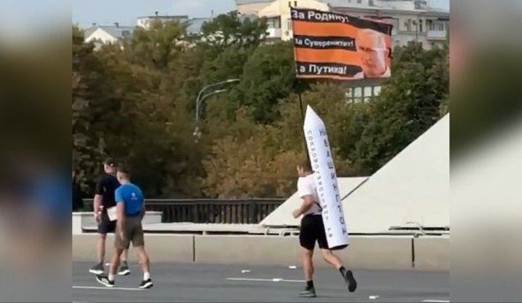 Россиянин бежит по российскому городу с флагом "За Родину! За суверенитет! За Путина" и макетом ракеты с надписью "На Вашингтон!" за спиной